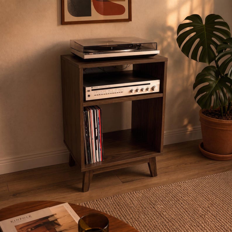 Puede incluir: Un soporte para tocadiscos de madera oscura con cuatro patas c&oacute;nicas, que cuenta con una superficie superior abierta para el tocadiscos, un estante central con un receptor de audio plateado y un compartimento inferior para discos.