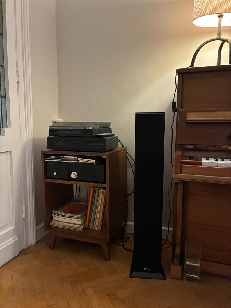Puede incluir: Un mueble de madera para equipos de audio con dos estantes, que sostiene un tocadiscos, un receptor y un reproductor de CD. El estante inferior contiene una pila de libros y discos. Un altavoz de pie negro y alto est&aacute; junto al mueble.