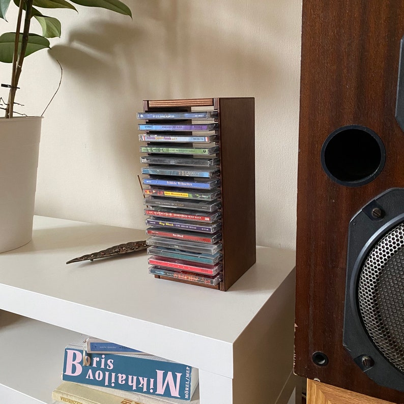 Peut inclure: Un porte-CD en bois avec une finition brun fonc&eacute; contient une collection de CD color&eacute;s. Le porte-CD est pos&eacute; sur une &eacute;tag&egrave;re blanche &agrave; c&ocirc;t&eacute; d'une grande enceinte brun fonc&eacute;.