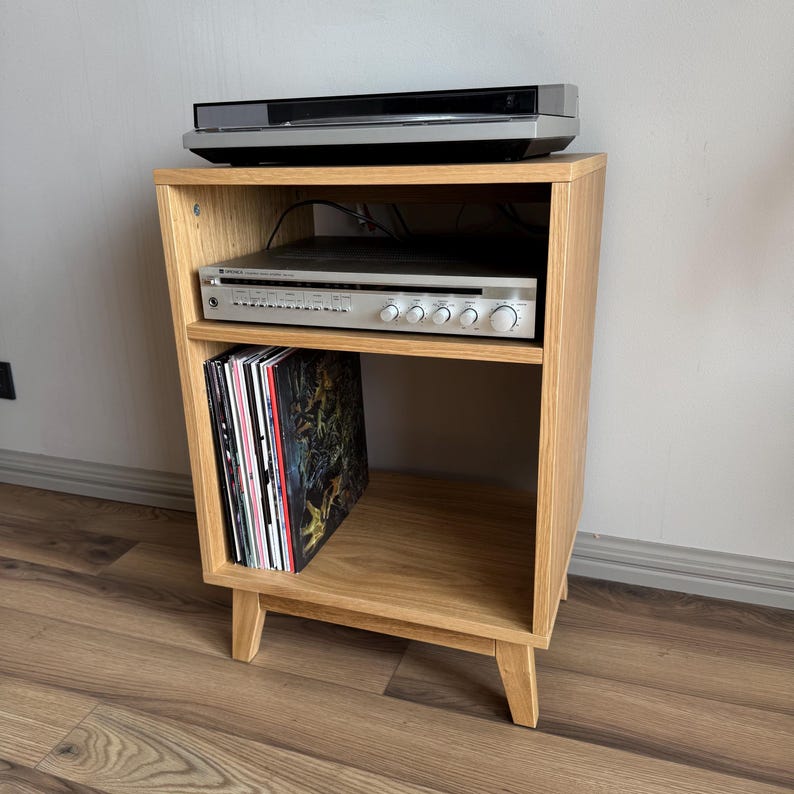 Puede incluir: Un soporte multimedia de madera de roble claro con cuatro patas c&oacute;nicas, que cuenta con un estante superior abierto con un tocadiscos negro, un estante central con un receptor de audio Sony plateado y discos de vinilo abajo.
