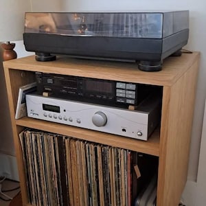 Puede incluir: Un mueble multimedia de madera clara con un tocadiscos negro en la parte superior. El estante central contiene un reproductor de CD Sony negro sobre un receptor de audio plateado. El estante inferior est&aacute; lleno de discos de vinilo.