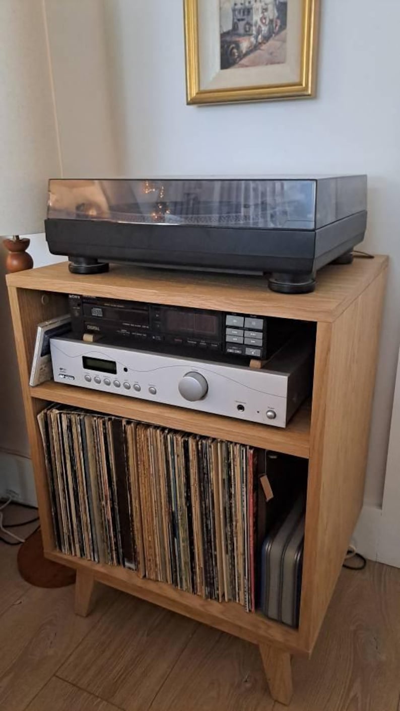 Puede incluir: Un mueble multimedia de madera clara con un tocadiscos negro en la parte superior. El estante central contiene un reproductor de CD Sony negro sobre un receptor de audio plateado. El estante inferior est&aacute; lleno de discos de vinilo.