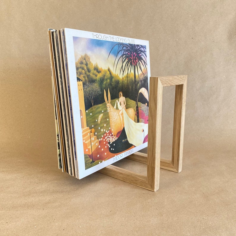 May include: A wooden record stand holding a stack of vinyl records. The record cover shows a painting of a woman and a white animal in a woodland scene. The record is titled "Through the Looking Glass" by "Mood Swings".