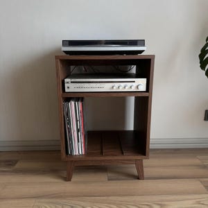 Puede incluir: Un mueble multimedia de madera oscura con cuatro patas c&oacute;nicas, que cuenta con un estante superior abierto con un tocadiscos negro, un estante central con un receptor de audio plateado y un estante inferior con discos de vinilo.