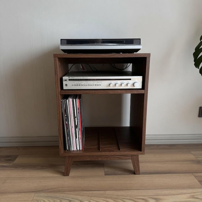 Puede incluir: Un mueble multimedia de madera oscura con cuatro patas c&oacute;nicas, que cuenta con un estante superior abierto con un tocadiscos negro, un estante central con un receptor de audio plateado y un estante inferior con discos de vinilo.