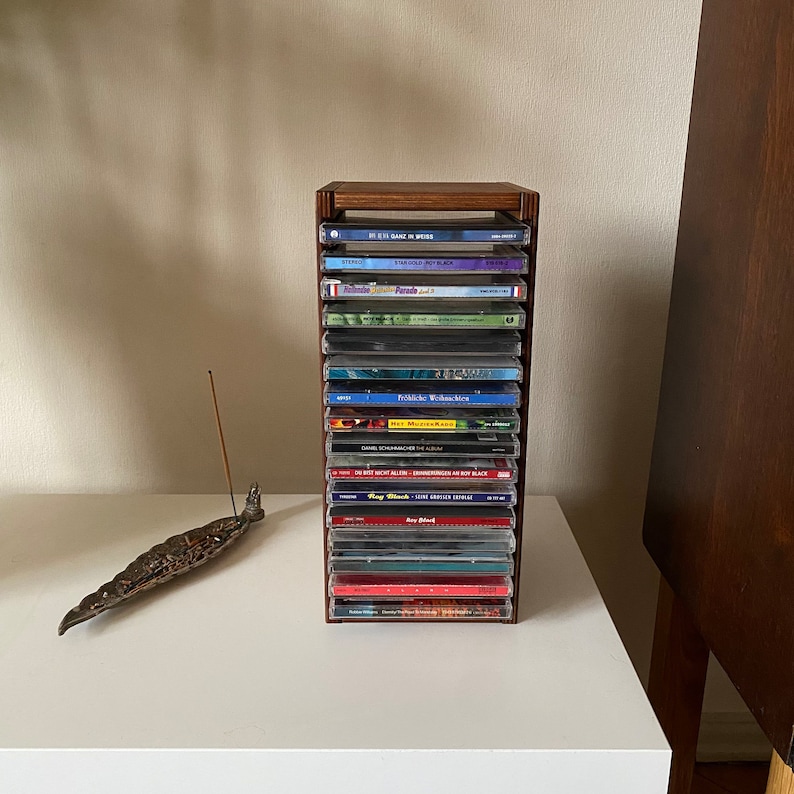 Peut inclure: Un porte-CD en bois avec 12 CD empil&eacute;s &agrave; l'int&eacute;rieur. Les CD ont des titres et des artistes vari&eacute;s. Le porte-CD est pos&eacute; sur une surface blanche.