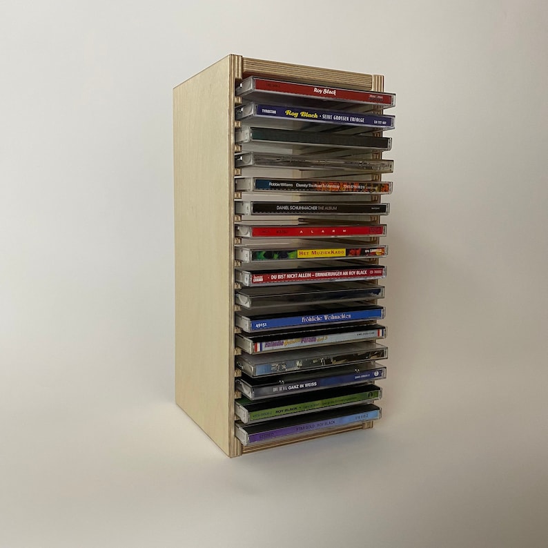 Peut inclure: Une tour de rangement en bois pour CD avec 18 fentes, contenant une vari&eacute;t&eacute; de CD avec diff&eacute;rents titres et artistes. La tour est en bois clair et a une finition naturelle.