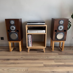 Puede incluir: Dos altavoces de madera oscura con tres controladores circulares cada uno, montados sobre soportes de madera clara. Entre ellos hay un mueble de audio de madera clara con un receptor plateado y discos de vinilo.