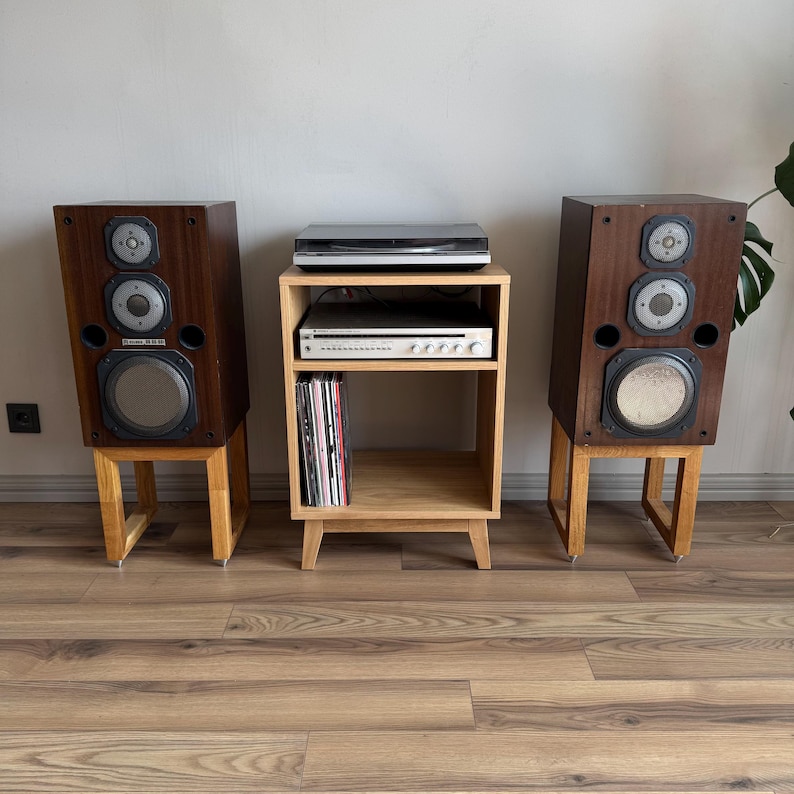 Puede incluir: Dos altavoces de madera oscura con tres controladores circulares cada uno, montados sobre soportes de madera clara. Entre ellos hay un mueble de audio de madera clara con un receptor plateado y discos de vinilo.