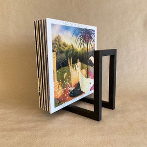 May include: A black wooden record stand holding a stack of vinyl records. The record cover features a painting of a woman and a white animal in a forest setting. The title of the record is "Through the Looking Glass" by "Moody Blues".
