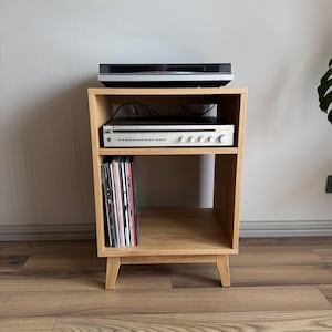 May include: A light oak wood media stand with four tapered legs, featuring an open top shelf holding a black turntable, a middle shelf with a silver audio receiver, and a bottom shelf containing a vertical row of vinyl records in colourful sleeves.