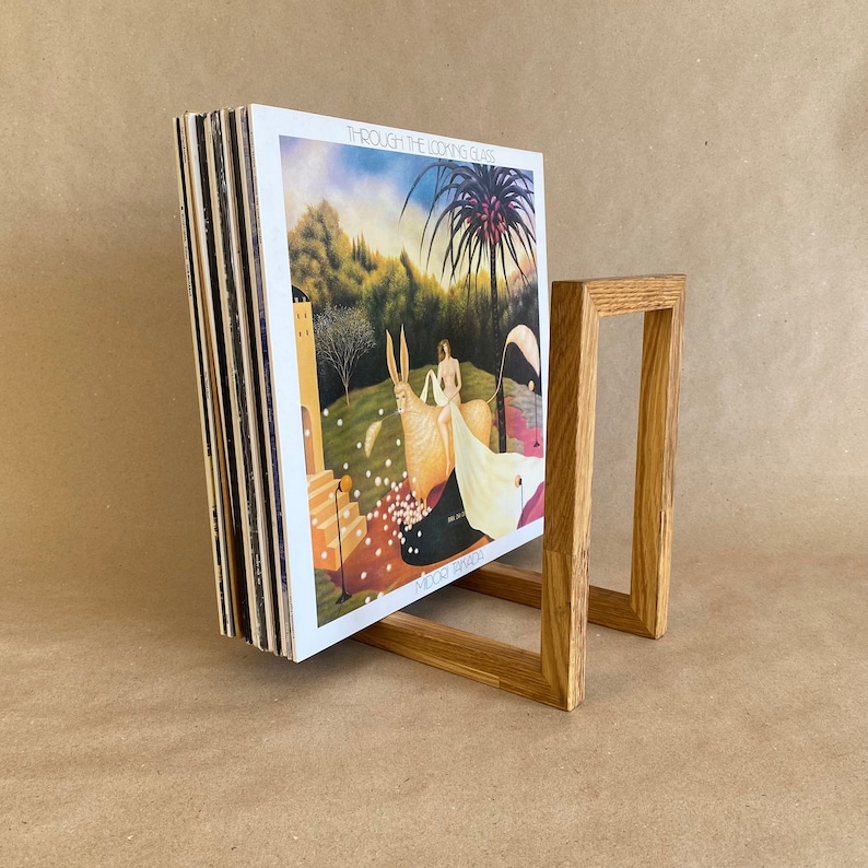 May include: A wooden record stand with a stack of vinyl records. The record cover features a painting of a white rabbit and a woman in a white dress. The album title is "Through the Looking Glass" by "Moriarty"