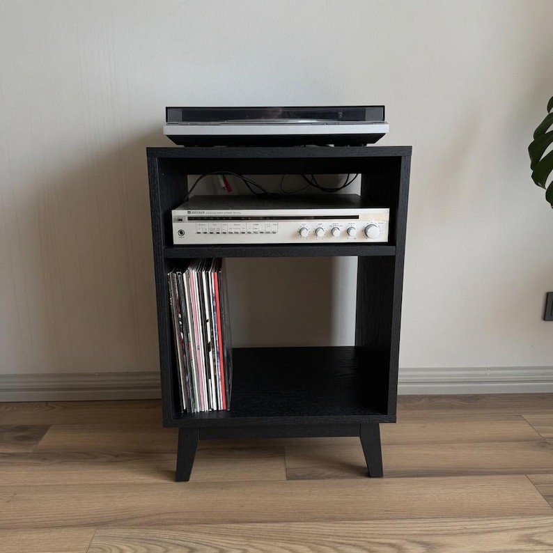 Puede incluir: Un soporte multimedia de madera negra con cuatro patas c&oacute;nicas, que cuenta con un estante superior abierto con un tocadiscos, un estante central con un receptor plateado y un estante inferior con discos de vinilo.