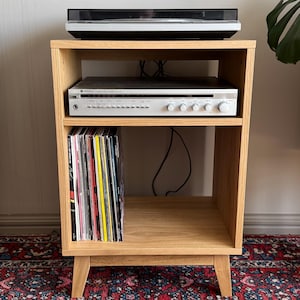 Puede incluir: Un mueble multimedia de madera de roble claro con cuatro patas c&oacute;nicas. La superficie superior sostiene un tocadiscos negro. El estante central contiene un amplificador integrado Optonica plateado. El estante inferior sostiene discos de vinilo.