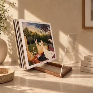 May include: A clear acrylic and dark wood vinyl record display stand holding a collection of records. The front record features the album cover Through the Looking Glass by Midori Takada, depicting a surreal landscape with a figure and a large animal.