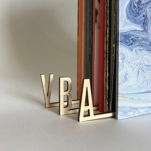 Peut inclure: Des serre-livres en bois en forme des lettres "VBA" dans une couleur brun clair. Les serre-livres maintiennent une pile de livres avec des dos colorés et une couverture marbrée bleu et blanc.