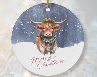 以下が含まれることがあります： ハイランド牛のイラストが描かれた丸いクリスマスオーナメント。牛は緑と青のスカーフ、リース、電飾を身につけています。「Merry Christmas」の文字が赤い筆記体で書かれています。