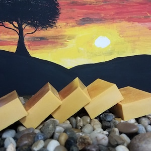 May include: Five bars of orange soap arranged in a pyramid shape on a bed of grey and brown stones. The soap is a bright orange colour and has a smooth, polished finish. The stones are various sizes and shapes, and they are arranged in a random pattern. The background is a painting of a sunset with a silhouette of a tree.