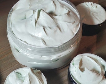 Puede incluir: Primer plano de un tarro de crema corporal batida de color verde claro. El tarro está lleno hasta el borde con el producto cremoso, y hay tres tarros más pequeños del mismo producto en el fondo.
