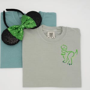 May include: Two t-shirts, one teal and one gray, are stacked. A black headband with sequined mouse ears and a green sequined bow sits on top of the teal shirt. The gray shirt has a green dinosaur outline.