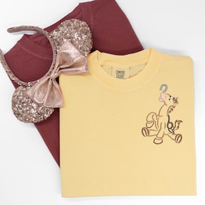 May include: A yellow Comfort Colors t-shirt with an embroidered cartoon character. A maroon t-shirt and a sequined Minnie Mouse ear headband with a pink bow are also in the image. The items are on a white background.