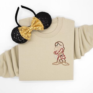 May include: A beige crewneck sweatshirt featuring an embroidered character with crossed arms, wearing a red tunic and brown boots. Beside the sweatshirt is a black headband with two round ears covered in black sequins and a gold sequined bow.
