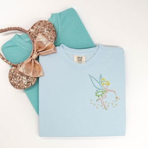 May include: A light blue crewneck sweatshirt featuring an embroidered Tinker Bell outline with a wand and stars, layered over a teal t-shirt. A pair of rose gold sequined mouse ears with a matching bow is positioned on the left side of the garments.
