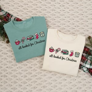 May include: A sage green sweatshirt and a cream sweatshirt, each featuring a graphic print of a mug, candy cane, stack of books, stocking, and candle, with the text "all booked for Christmas" written in black cursive lettering below the icons.