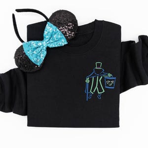 May include: A black long-sleeve sweatshirt featuring an embroidered neon-style graphic of a figure in a top hat and cape holding a bucket with a face on it. Beside the sweatshirt is a black headband with two round ears and a bright blue sequined bow.