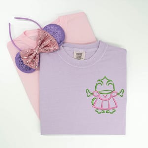 May include: A light purple t-shirt featuring an embroidered green and pink frog character on the chest, layered over a light pink t-shirt. A pair of purple sequined ears with a pink sequined bow is placed on top of the folded shirts.