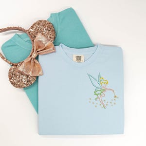 May include: A light blue crewneck sweatshirt featuring an embroidered Tinker Bell outline on the chest, paired with a teal t-shirt and a pair of rose gold sequined mouse ears with a matching fabric bow. The sweatshirt has a Comfort Colors label.