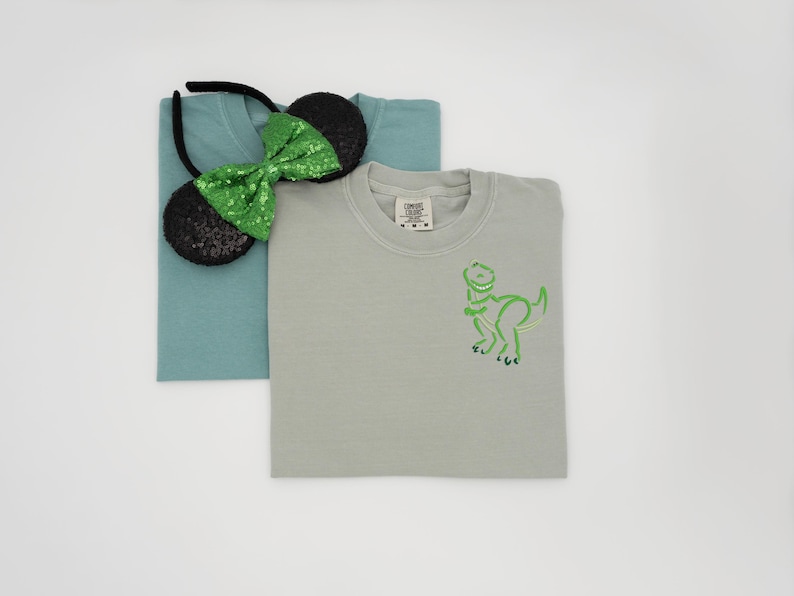 May include: Two t-shirts, one teal and one gray, are stacked. A black headband with sequined mouse ears and a green sequined bow sits on top of the teal shirt. The gray shirt has a green dinosaur outline.