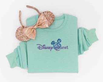 May include: A mint green long-sleeve sweatshirt featuring the embroidered purple text Disney Quest with a stylized circular icon above the word Quest. Resting on the sweatshirt is a pair of rose gold sequined headband ears with a bow.