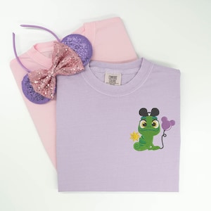 May include: A light purple t-shirt featuring an embroidered green chameleon character holding a purple balloon and wearing black mouse ears, with a small yellow sun icon. A light pink t-shirt and purple sequined mouse ear headband are also visible.