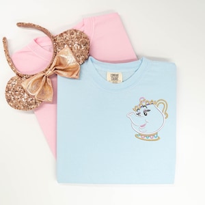 May include: A light blue t-shirt featuring an embroidered graphic of a teapot character on the chest, layered over a plain pink t-shirt. Beside the shirts is a pair of rose gold sequined ear headbands with a matching metallic bow.