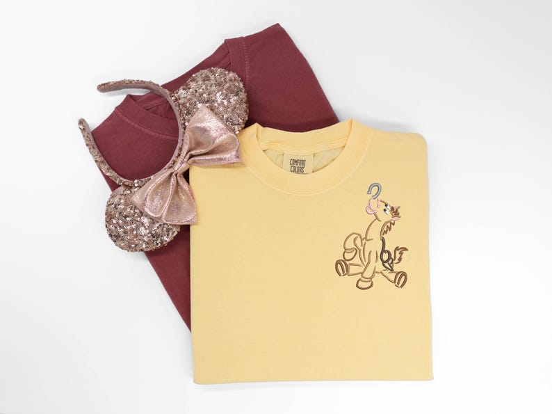May include: A yellow Comfort Colors t-shirt with an embroidered cartoon character. A maroon t-shirt and a sequined Minnie Mouse ear headband with a pink bow are also in the image. The items are on a white background.