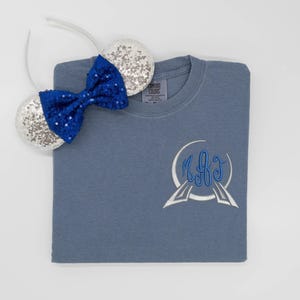 May include: A slate blue t-shirt featuring a white and blue embroidered monogram on the chest, paired with a headband featuring two silver sequined ears and a bright blue sequined bow. The shirt has a Comfort Colors tag at the inner neckline.