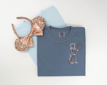 May include: A slate blue t-shirt featuring an embroidered rat wearing a chef hat and holding a large wooden spoon, layered over a light blue t-shirt and a pair of rose gold sequined mouse ears with a matching bow attached to a headband.