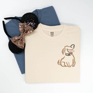 May include: A cream-colored crewneck sweatshirt featuring an embroidered brown dog outline on the chest, layered over a blue t-shirt and a black headband with sequined ears and a bronze bow. The sweatshirt has a Comfort Colors brand label.