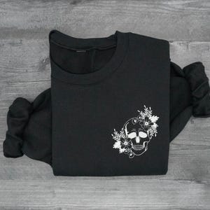 May include: Black sweatshirt featuring a white floral skull design on the front. The crew neck sweatshirt is made of a soft material and is folded on a gray wooden surface. The design includes a skull with flowers.