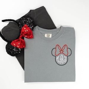 May include: A gray t-shirt with a Minnie Mouse design and a pair of black sequined ears with a red sequined bow. The Minnie Mouse design features a red bow and a monogram.