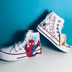 May include: White high-top sneakers with a hand-painted design featuring Spiderman, a web, and the name "STEFANO".