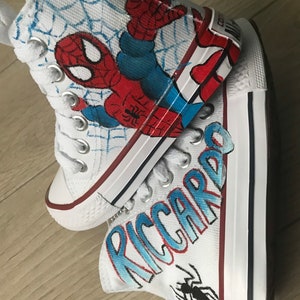 May include: A pair of white high-top canvas shoes with a hand-painted design. One shoe features a Spiderman design with red, blue, and black colors. The other shoe has the name "RICCARDO" written in blue and red with a black spider design.