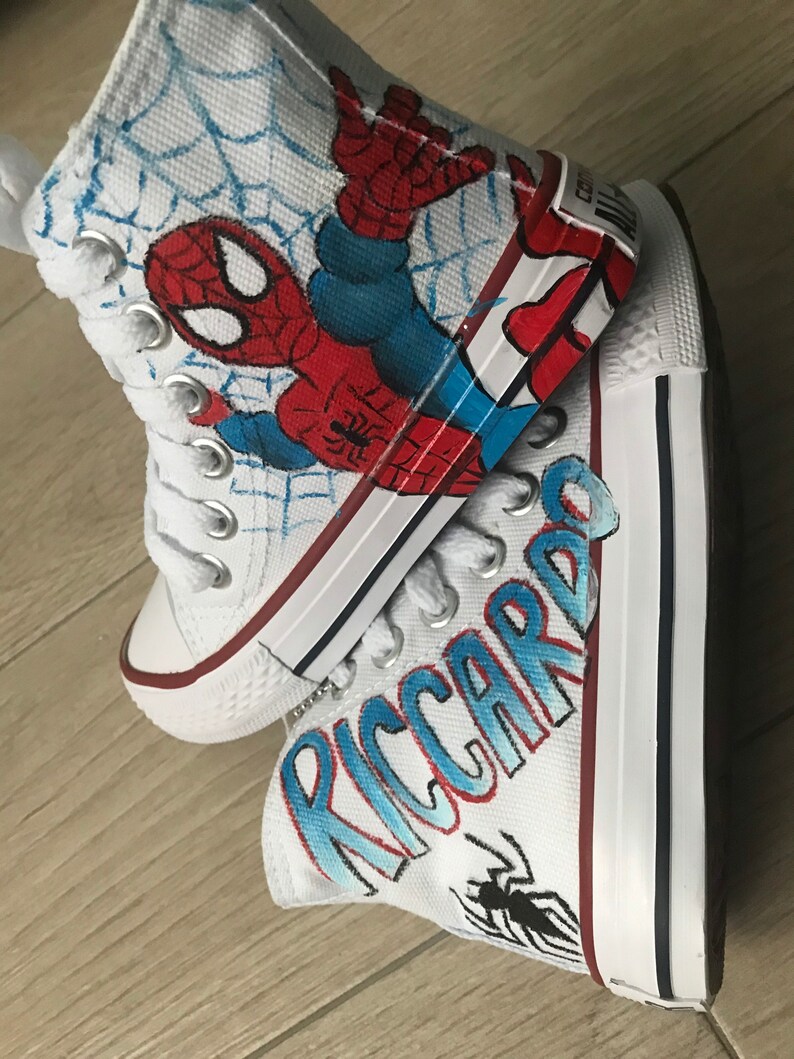 May include: A pair of white high-top canvas shoes with a hand-painted design. One shoe features a Spiderman design with red, blue, and black colors. The other shoe has the name "RICCARDO" written in blue and red with a black spider design.