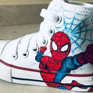 May include: White high-top sneakers with a hand-painted design of Spiderman in red, blue, and black. The design includes a web pattern in blue.
