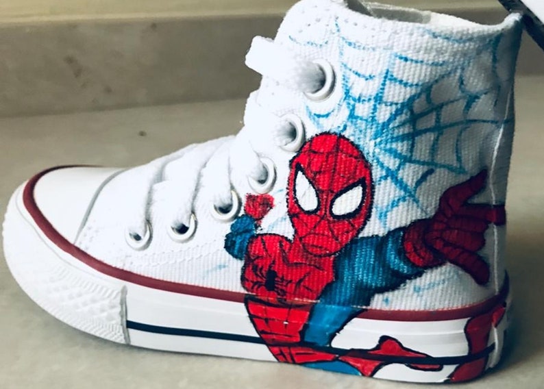 May include: White high-top sneakers with a hand-painted design of Spiderman in red, blue, and black. The design includes a web pattern in blue.