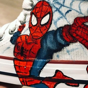 May include: White canvas high-top sneakers with a hand-painted design of Spider-Man in red and blue. The design features Spider-Man's face and a web pattern.