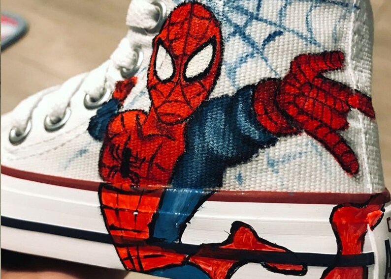 May include: White canvas high-top sneakers with a hand-painted design of Spider-Man in red and blue. The design features Spider-Man's face and a web pattern.