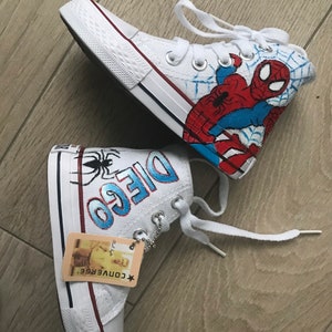 May include: White high-top canvas shoes with a red and blue Spiderman design on one shoe and the name "Diego" written in blue on the other shoe. The shoes have white laces and a red Converse logo.