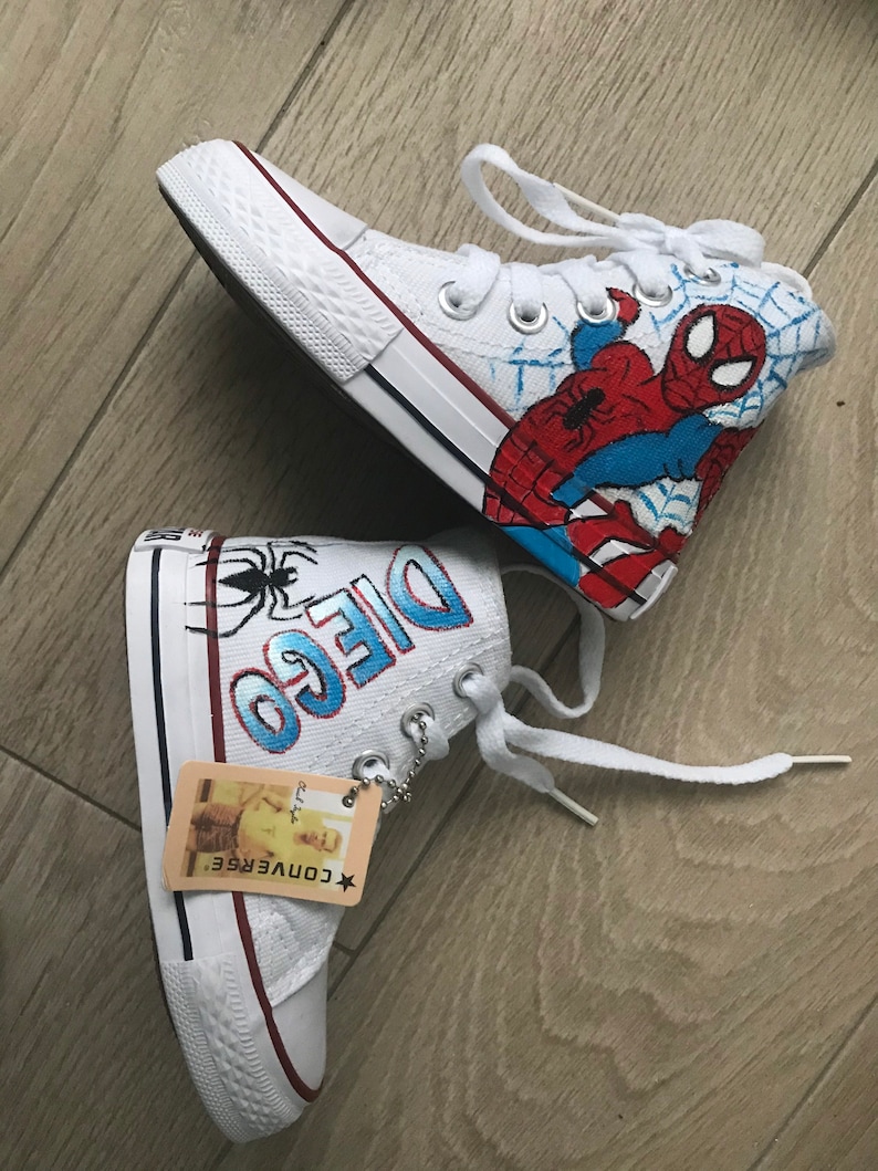 May include: White high-top canvas shoes with a red and blue Spiderman design on one shoe and the name "Diego" written in blue on the other shoe. The shoes have white laces and a red Converse logo.